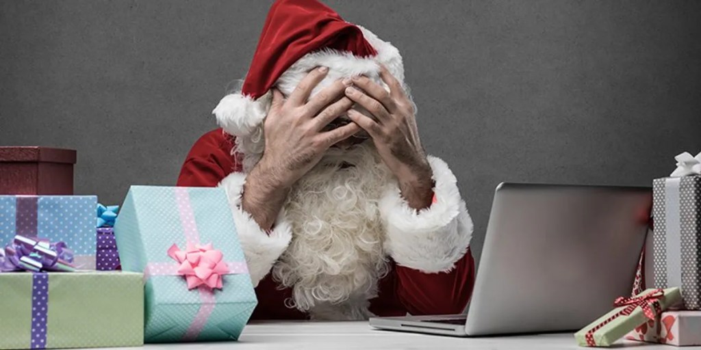 Santa Claus looking stressed while sitting at a desk with his hands on his head, surrounded by colorful Christmas gifts and a laptop.