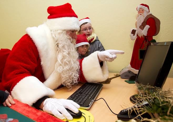 Santa Claus sitting at a desk, pointing at a computer screen with two children watching him, festive decorations in the background.
